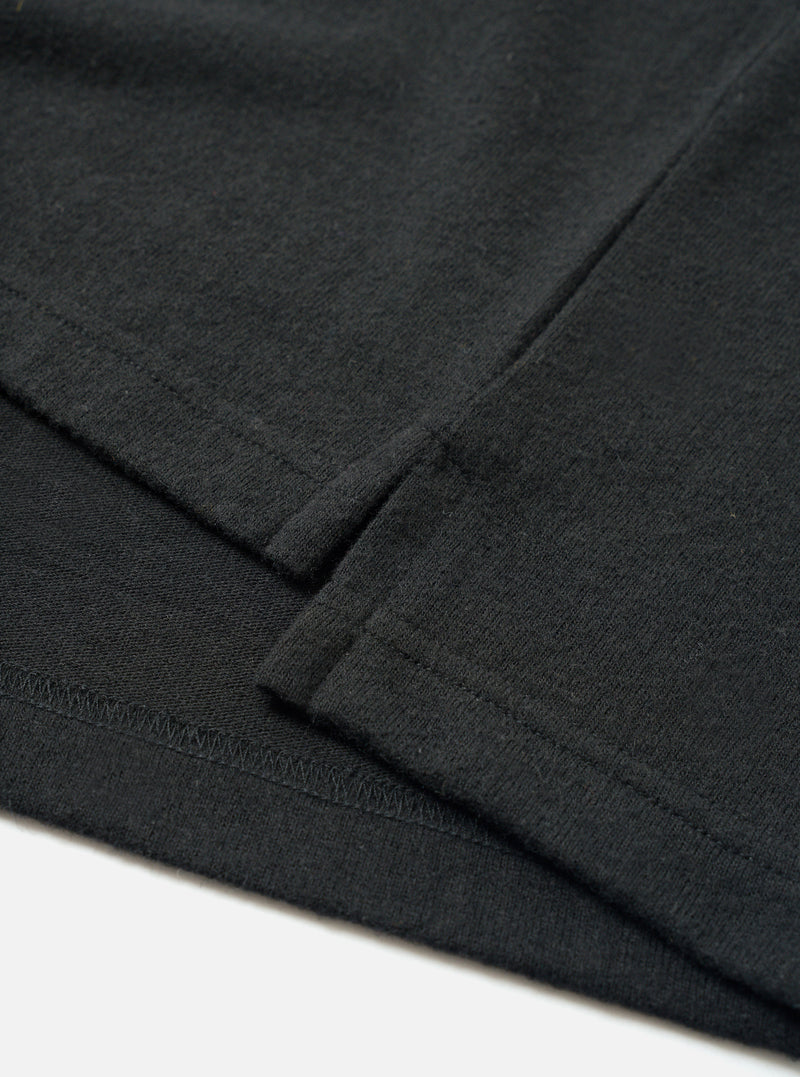 Close-up flat lay detail of the black recycled wool mix long-sleeve pocket tee. The shot focuses on the chest patch pocket and sleeve cuff, showing the folded ribbed cuff edge with visible topstitching and a zigzag overlock seam. The fine-knit jersey fabric has a subtle heathered texture. White surface background.