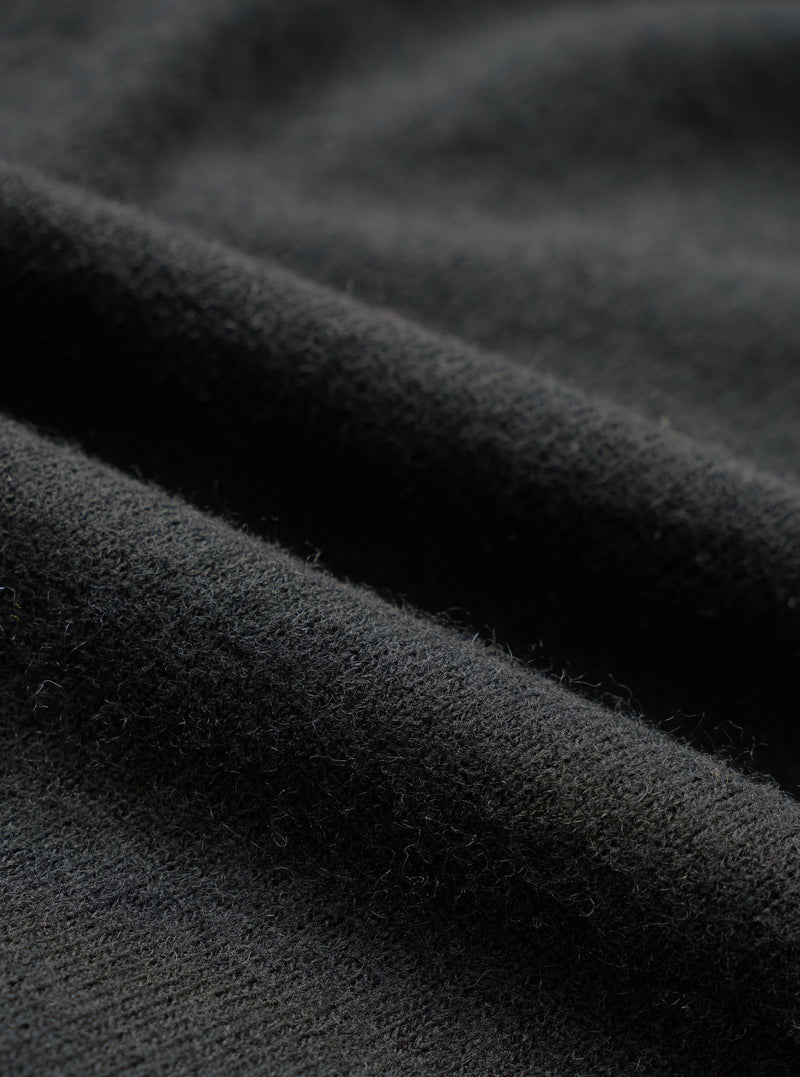 Close-up detail shot of the black recycled wool mix fabric, showing a soft, lightly brushed texture with fine fibres visible on the surface. The material has a dense, matte finish with gentle folds catching the light to reveal subtle tonal variation across the dark charcoal-black cloth.