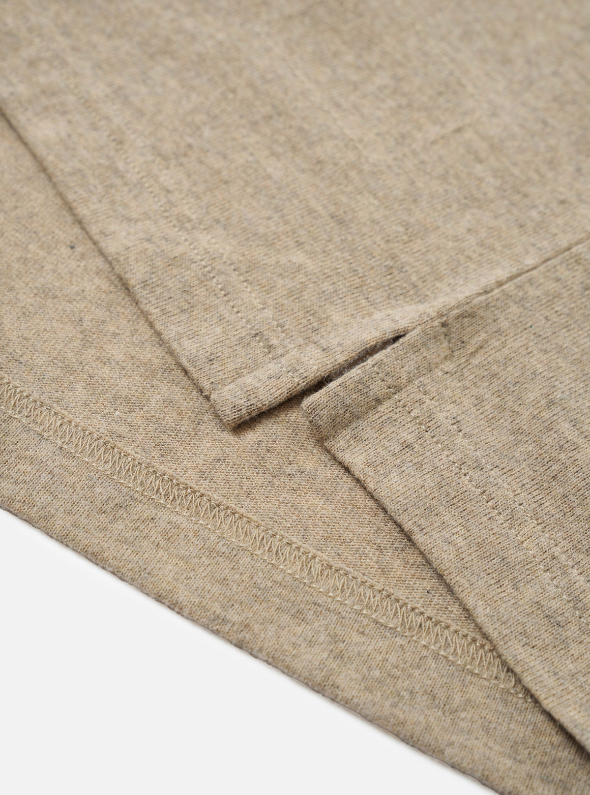 Close-up detail of a dark sand recycled wool mix long-sleeve pocket tee, flat on a white surface. The shot focuses on the curved hem with visible overlock stitching and a small side seam vent. The marl-textured fabric shows a heathered oatmeal tone with flecks of darker fibre throughout.