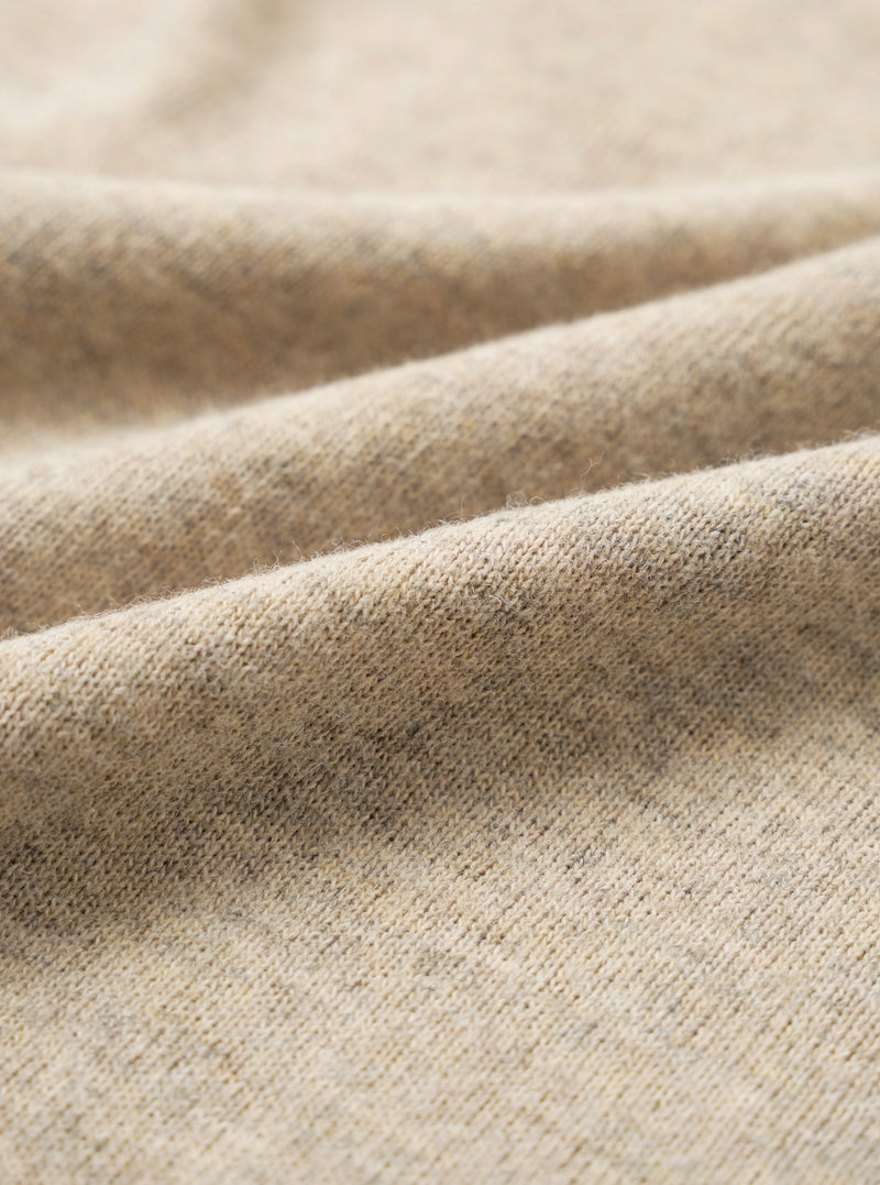 Close-up of dark sand recycled wool mix fabric, showing a fine knit texture with a soft, slightly haired surface. The oatmeal-toned yarn has subtle flecks of grey and brown throughout. Gentle folds run diagonally across the frame, highlighting the fabric's drape and weight.