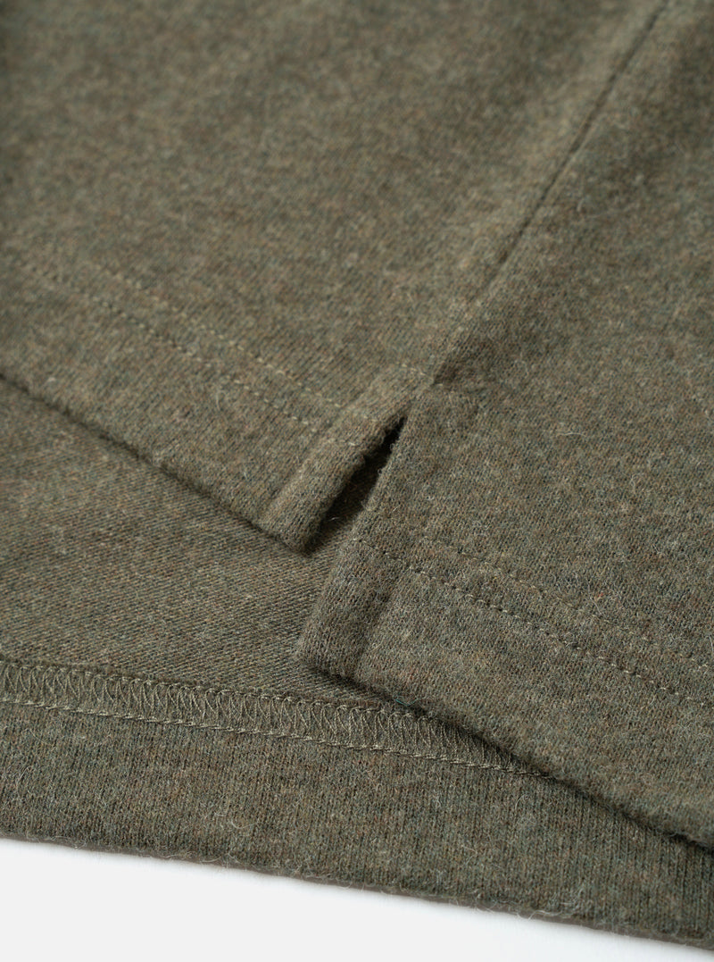 Close-up flat lay of an olive marl recycled wool mix long-sleeve pocket tee on a white surface. The shot focuses on the lower hem and cuff, showing a ribbed hem with overlock stitching, a folded ribbed cuff, and a small welt thumb loop detail. The heathered fabric has a soft, lightly textured finish.