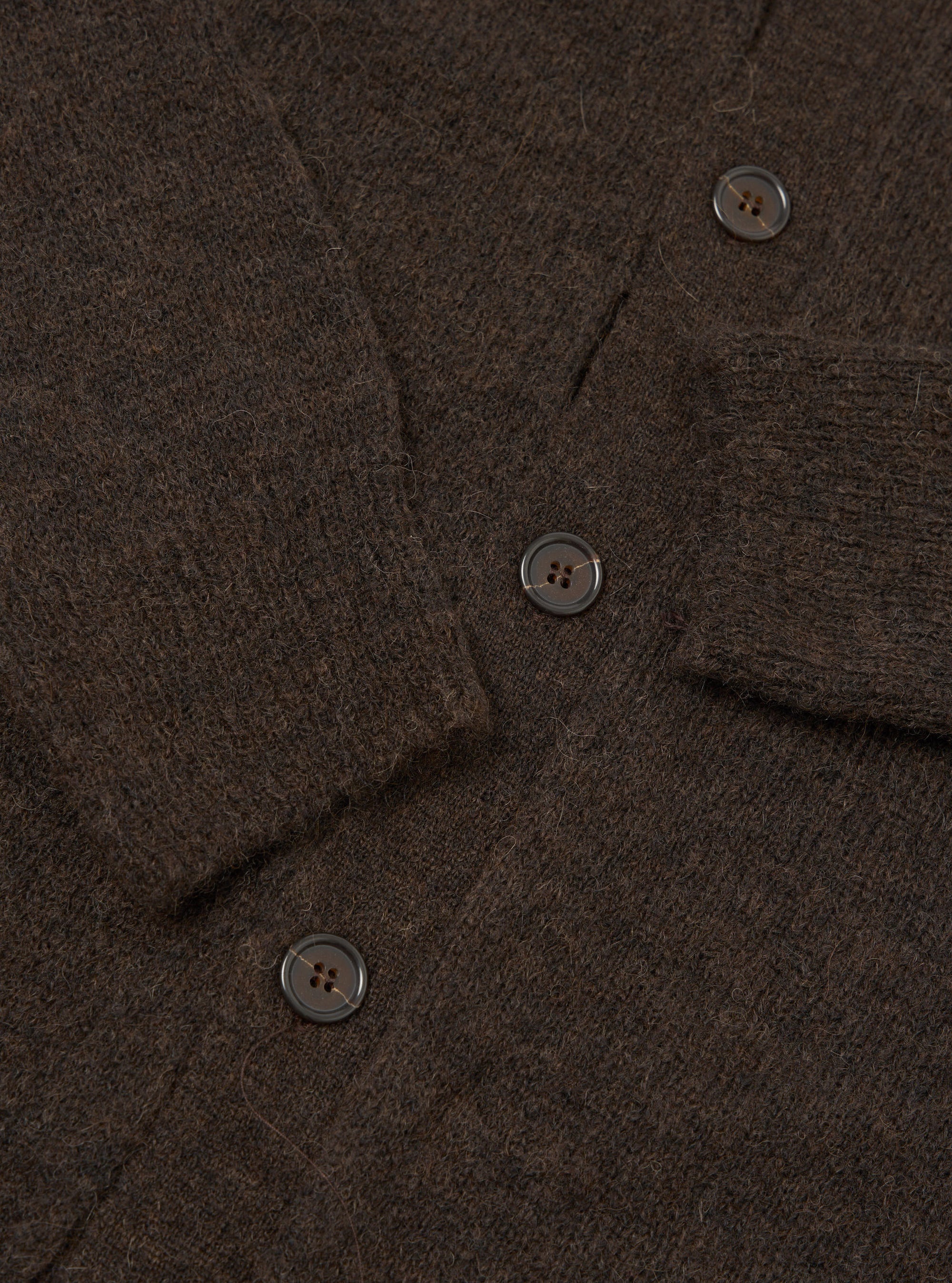 Close-up flat lay of the brown alpaca knit cardigan showing the button placket with three visible dark horn-effect four-hole buttons. The fluffy, hairy texture of the alpaca knit is prominent throughout. A ribbed front band runs along the placket edge, with the sleeve visible in the upper right corner.