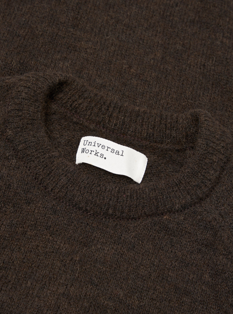 Close-up flat lay of a brown alpaca knit crew neck sweater. The ribbed crew neckline is visible at the top of the frame, with a white woven Universal Works brand label stitched inside. The fluffy, hairy texture of the alpaca knit fabric fills the frame. Shot on a plain surface.