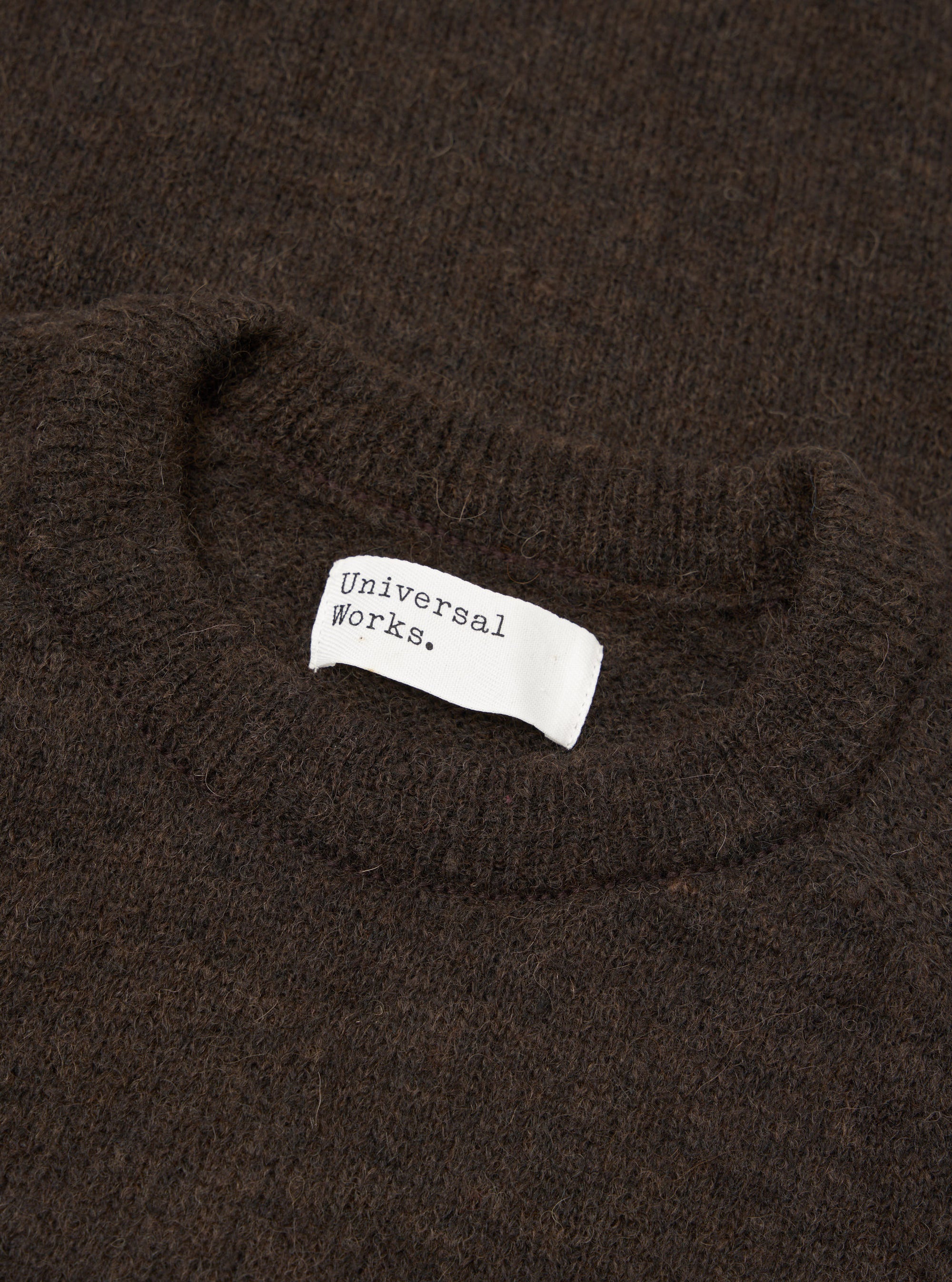 Close-up flat lay of a brown alpaca knit crew neck sweater. The ribbed crew neckline is visible at the top of the frame, with a white woven Universal Works brand label stitched inside. The fluffy, hairy texture of the alpaca knit fabric fills the frame. Shot on a plain surface.