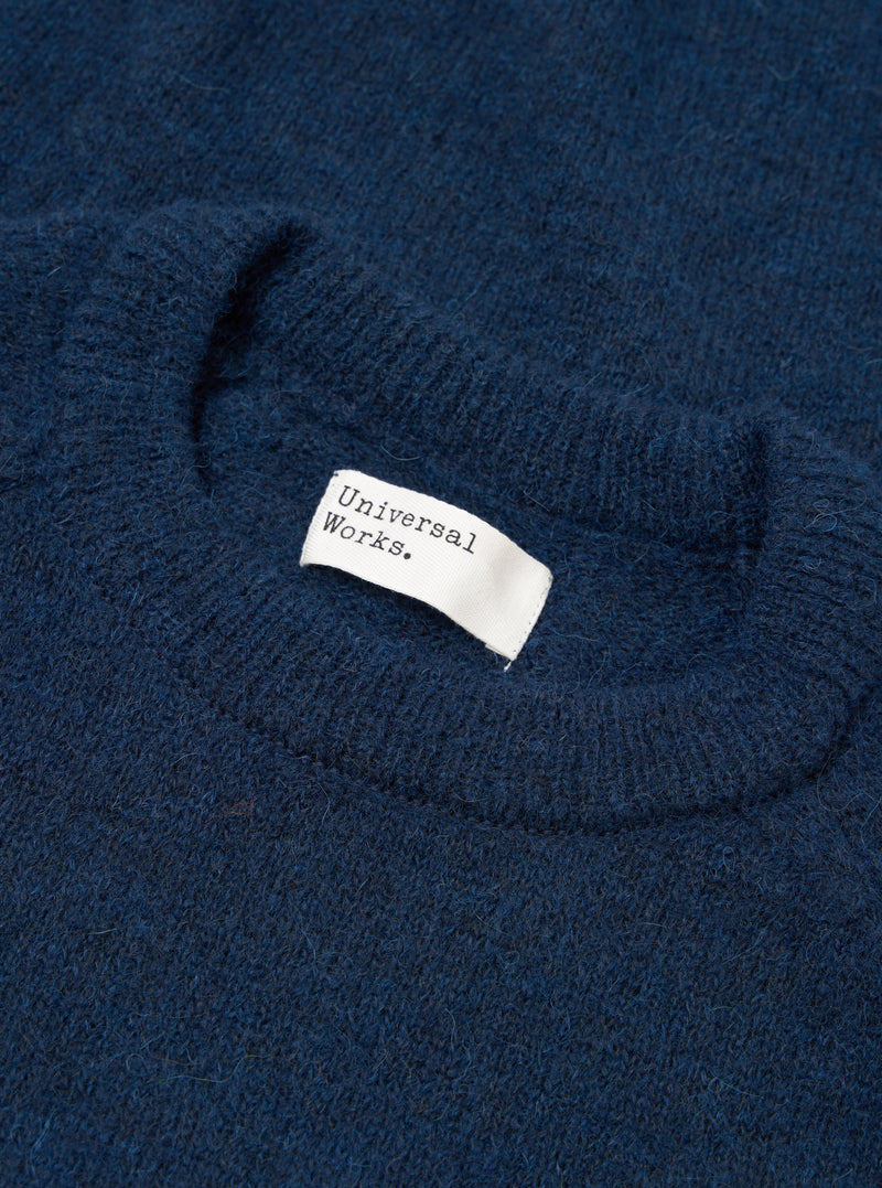 Close-up flat lay of the crew neck collar on a navy alpaca knit jumper. The ribbed neckline has a soft, fluffy texture. A white woven Universal Works label is visible inside the collar. The fabric shows a marl-flecked deep navy tone throughout.