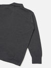 Close-up flat lay of a charcoal merino long-sleeve pullover knit shirt on a light grey seamless backdrop. The shot shows the upper back, shoulder and full length of one sleeve. A spread collar is visible at the top, the fabric has a fine knit texture, and a ribbed cuff finishes the sleeve. A partial hem vent is visible at the lower left.