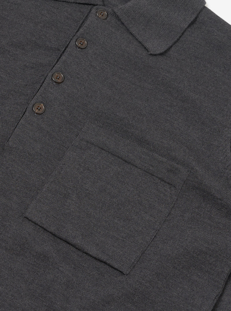 Close-up flat lay of a charcoal merino long-sleeve knit polo shirt on a white surface. The fine-knit fabric has a smooth, dense texture. Visible details include a spread collar, a four-button placket with dark tonal buttons, and a large patch chest pocket with a horizontal seam detail at the base.