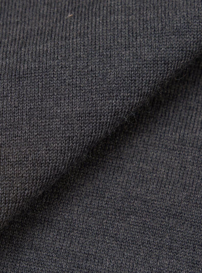 Close-up of the charcoal merino knit fabric, showing a fine, uniform jersey stitch with a soft, slightly textured surface. The dark grey tone has a subtle heathered quality. The fabric is folded to display drape and thickness against a plain white background.