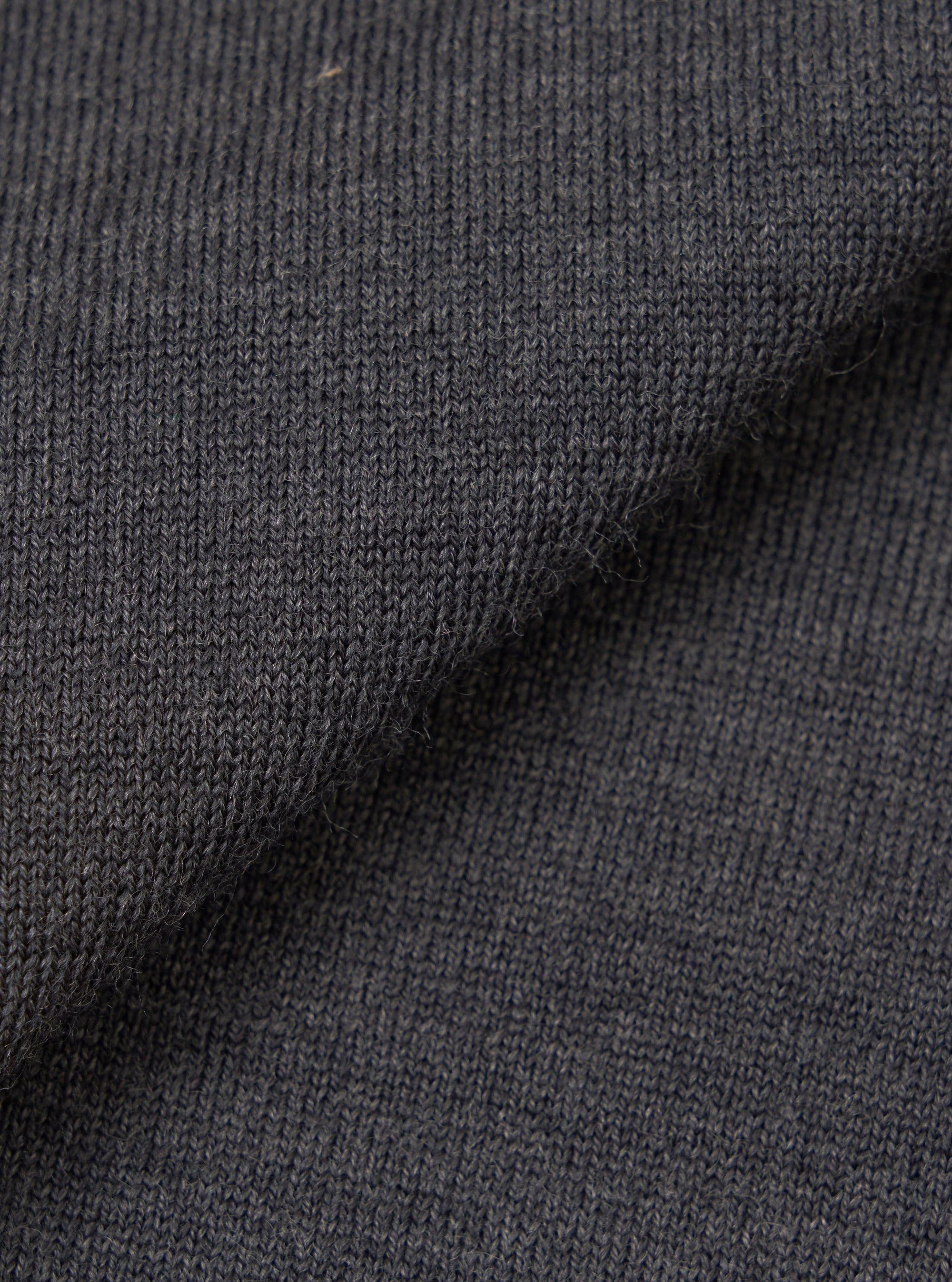 Close-up of the charcoal merino knit fabric, showing a fine, uniform jersey stitch with a soft, slightly textured surface. The dark grey tone has a subtle heathered quality. The fabric is folded to display drape and thickness against a plain white background.