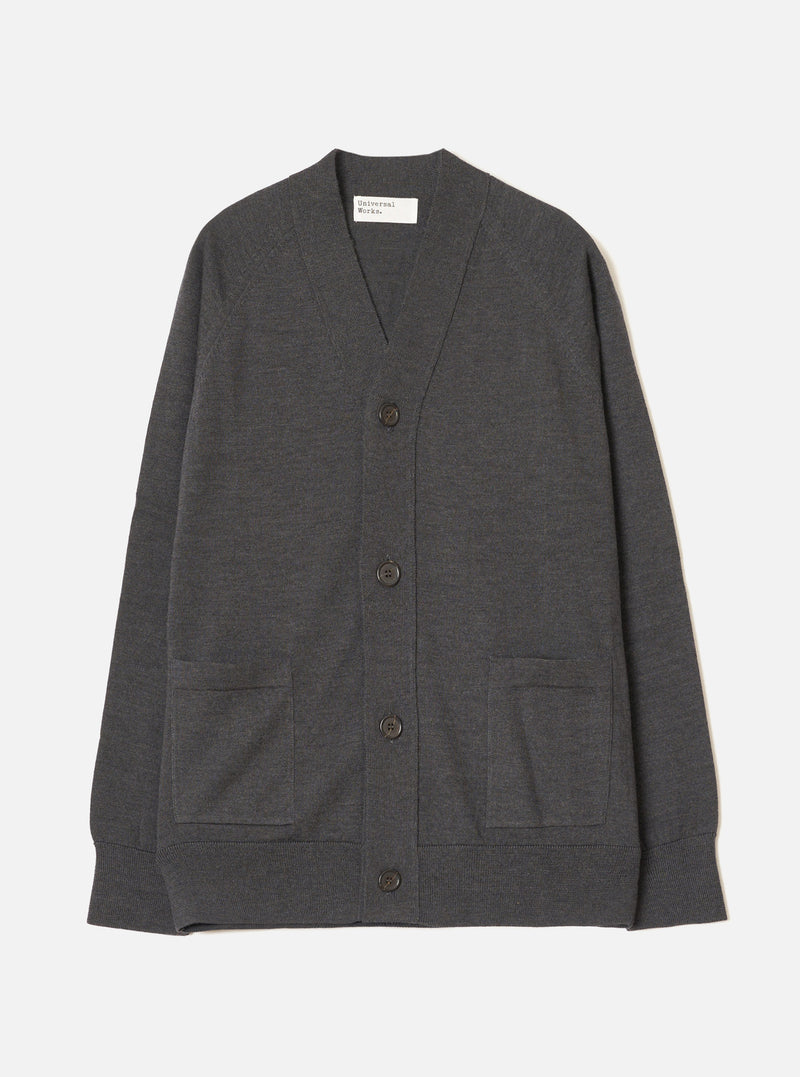 Charcoal merino cardigan laid flat on a light grey seamless backdrop. V-neckline with a woven Universal Works label inside. Four dark horn-effect buttons run down the front placket. Two patch pockets sit at the lower front. Ribbed cuffs and hem. Fine, smooth knit texture throughout. Full length, unbutoned view from the front.