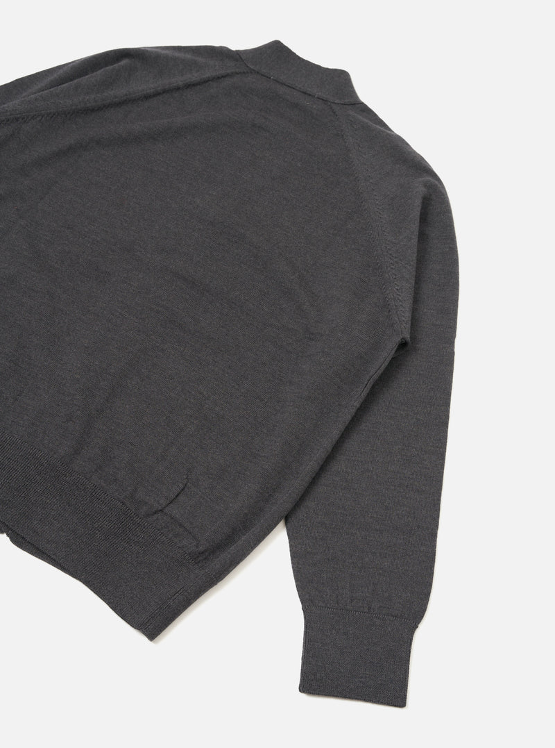 Flat lay of a charcoal merino loose cardigan on a white studio backdrop, shot from above at an angle. The fine-knit body shows a mock neck or collar at the top, a ribbed hem, and one full sleeve with a ribbed cuff. A seam detail runs along the sleeve join.