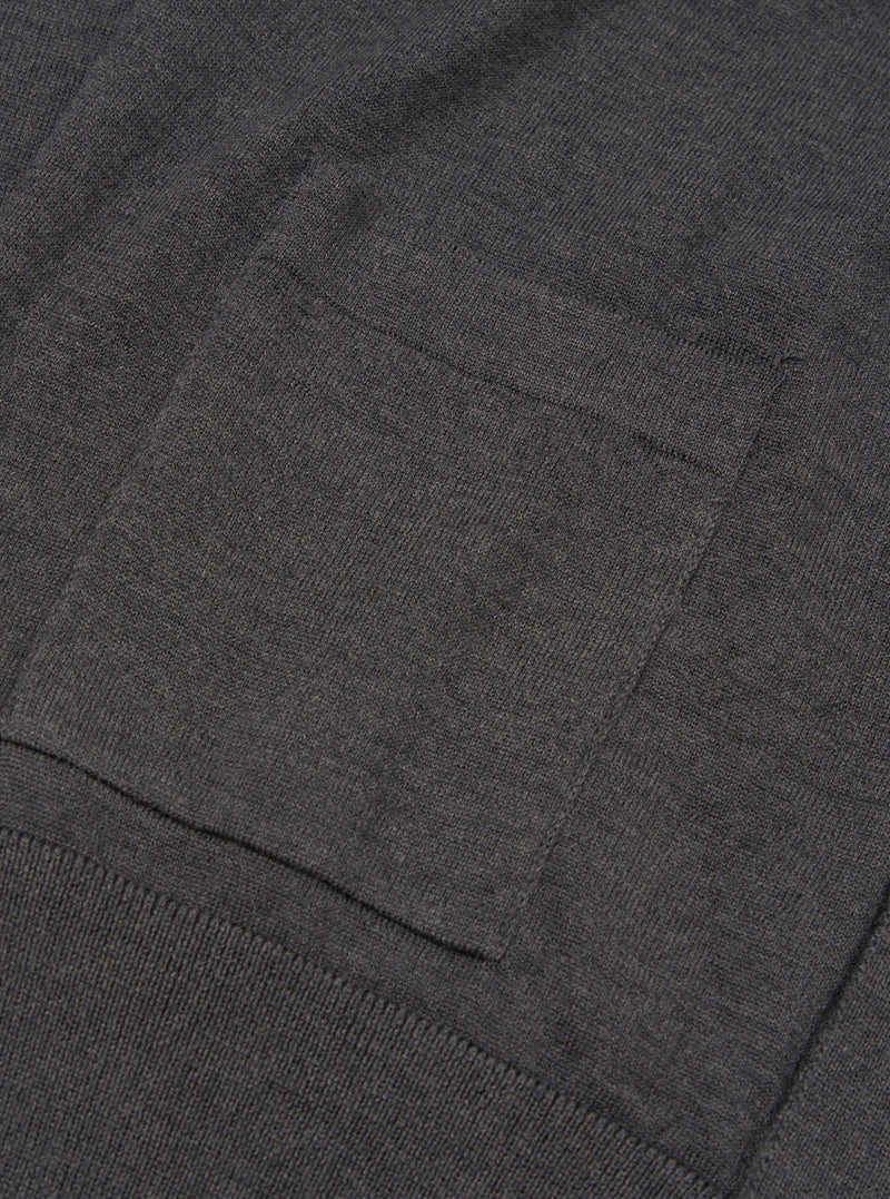 Close-up detail of a charcoal merino cardigan showing a patch chest pocket with neat stitched edges and a ribbed hem band below. The fine-knit merino fabric has a smooth, dense texture in a dark grey marl. Shot flat against a plain background.