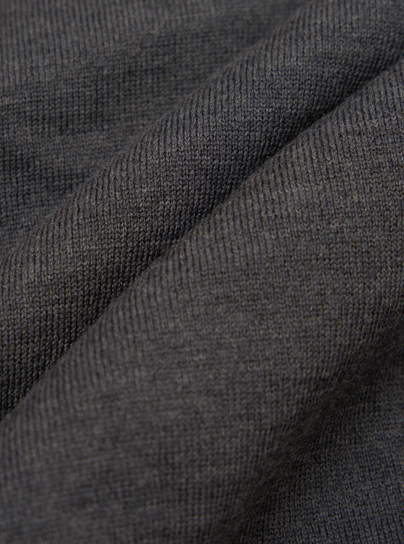 Close-up of charcoal merino knit fabric, showing a fine, even stocking stitch with a soft, slightly heathered texture. The fabric is gathered into soft folds, highlighting the drape and weight of the knit. No background is visible.
