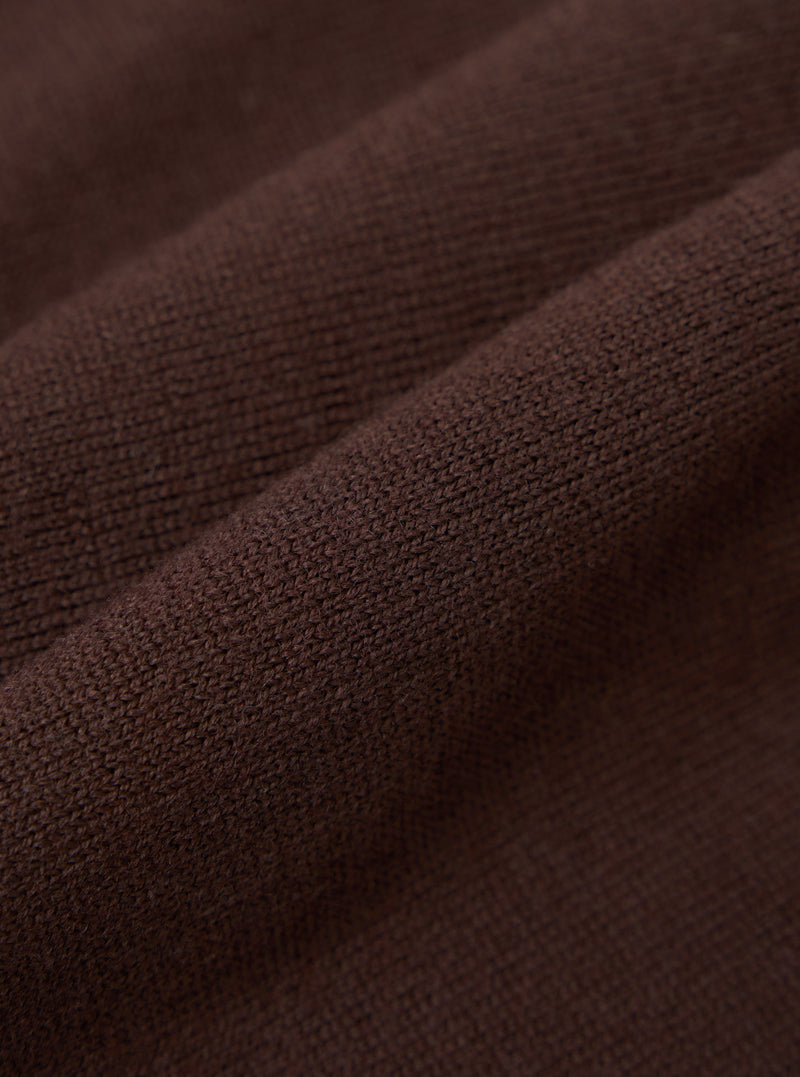 Close-up detail shot of the espresso brown merino knit fabric, showing the fine, even knit structure and a raised ribbed trim running diagonally across the frame. The dense, smooth texture and yarn quality are clearly visible against the draped folds of the garment.
