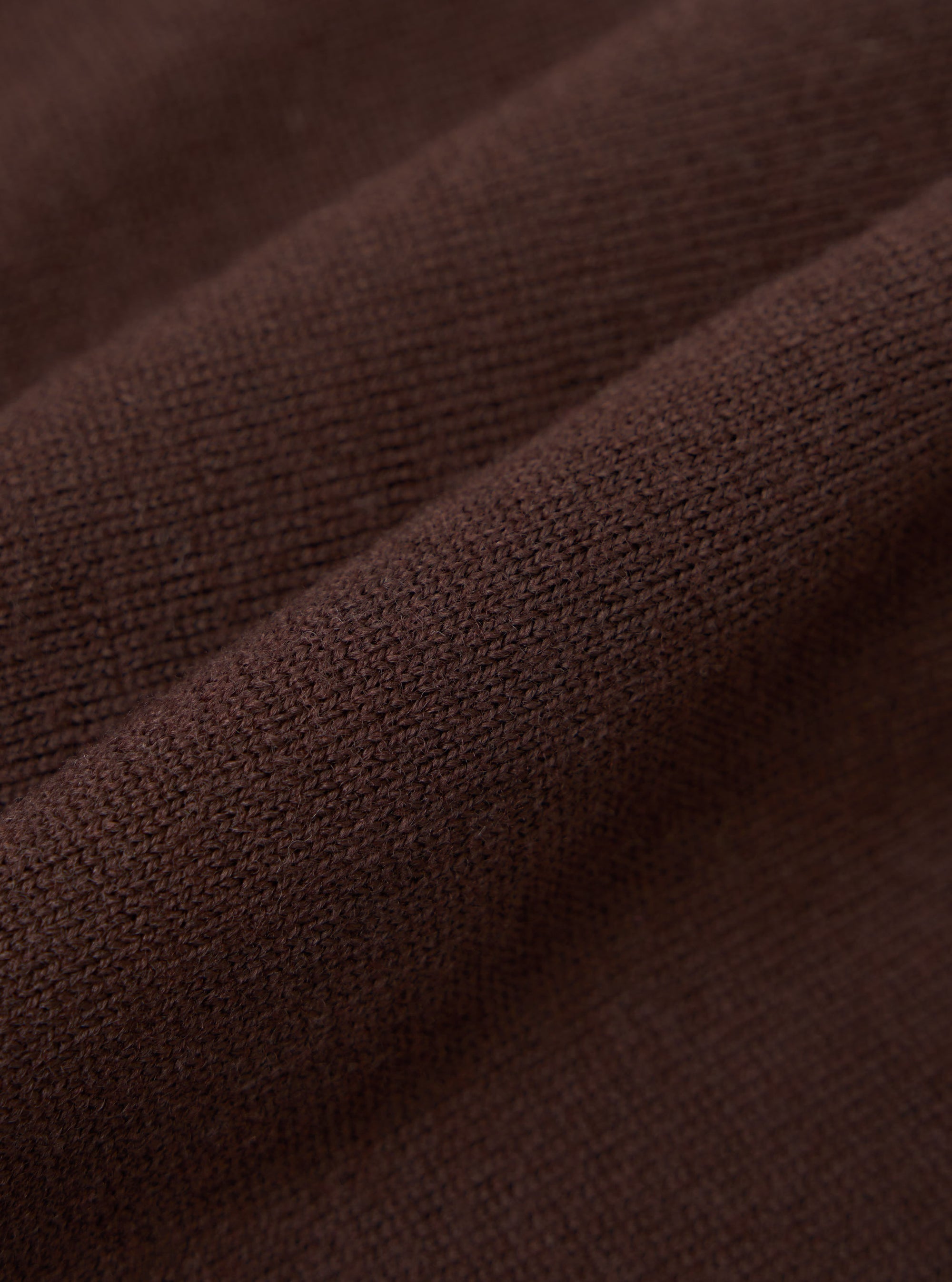 Close-up detail shot of the espresso brown merino knit fabric, showing the fine, even knit structure and a raised ribbed trim running diagonally across the frame. The dense, smooth texture and yarn quality are clearly visible against the draped folds of the garment.