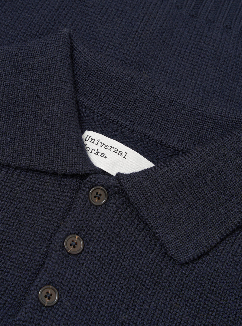 Close-up flat lay of a navy Shetland knit sweater showing the collar and button placket. The textured knit fabric is visible throughout. A white Universal Works woven label sits inside the collar. Three dark horn-effect buttons are visible on the placket. Shot on a plain white studio backdrop.