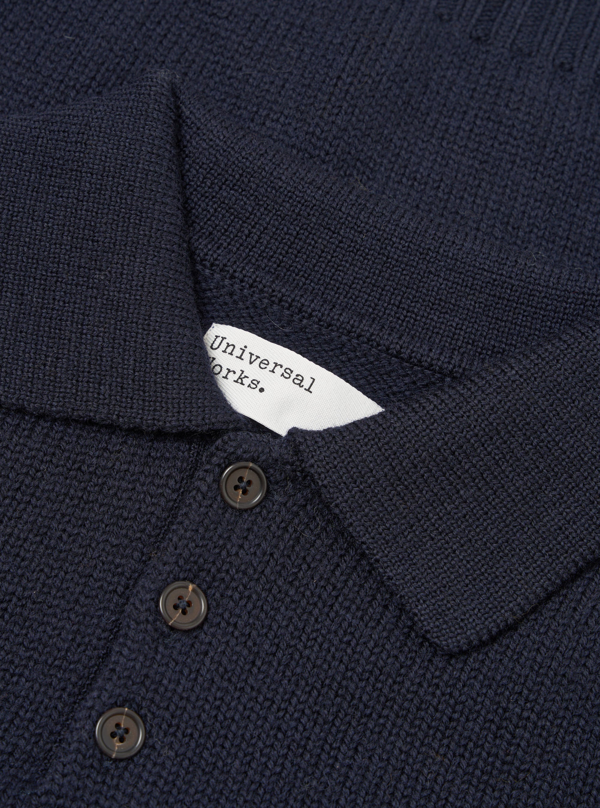 Close-up flat lay of a navy Shetland knit sweater showing the collar and button placket. The textured knit fabric is visible throughout. A white Universal Works woven label sits inside the collar. Three dark horn-effect buttons are visible on the placket. Shot on a plain white studio backdrop.