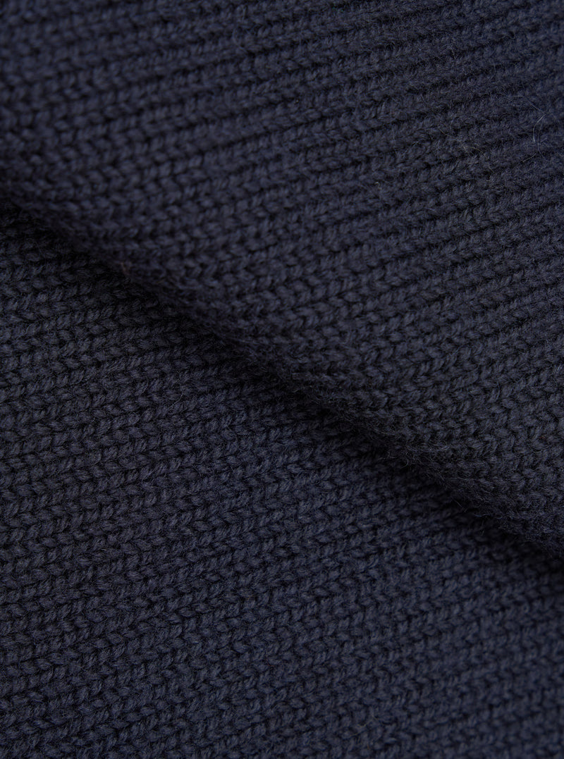 Close-up detail shot of the navy Shetland Wild Curtis sweater fabric. The chunky knit texture is shown in high detail, revealing a moss or seed stitch construction with a ribbed section visible at the top. The wool yarn is a deep navy, shot against no background.