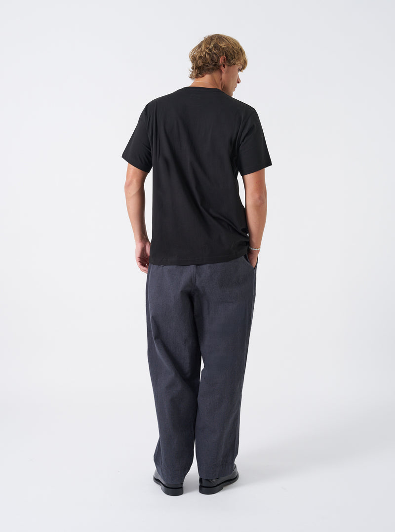 A model wearing the Grey Liverpool Check Oxford Pant, shown from the side against a neutral white backdrop. The photo highlights the comfortable fit, pleated waistband, side pockets, and full length drape of the checked trousers.