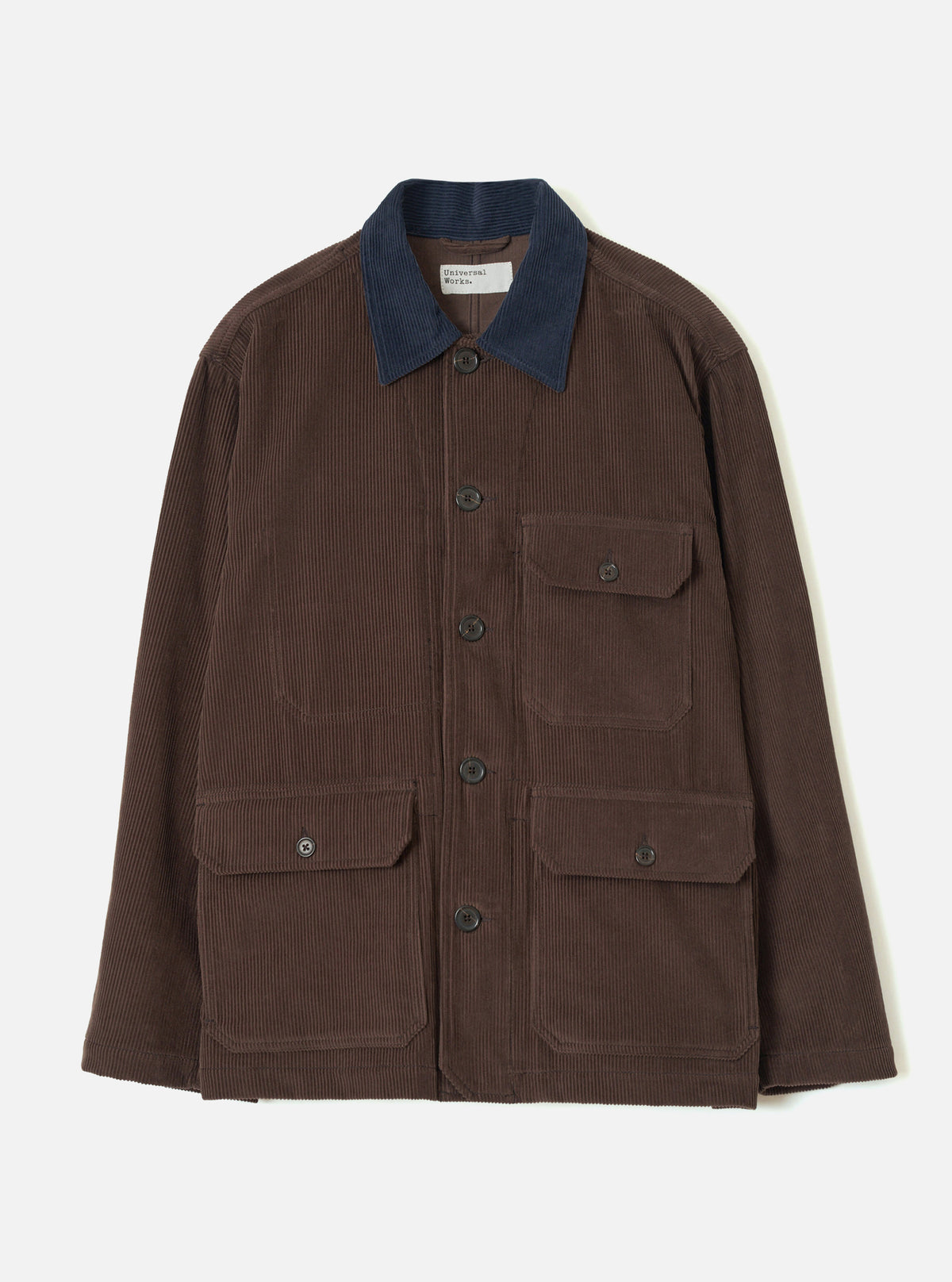 Cacao brown corduroy utility jacket laid flat on a light grey backdrop. Features a contrast navy corduroy collar, five dark buttons down the front placket, one buttoned chest patch pocket, and two large buttoned patch pockets at the hip. A Universal Works woven label sits inside the collar.