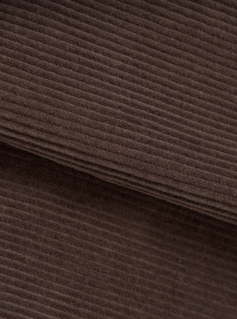 Close-up of the cacao brown corduroy fabric of the utility jacket, showing wide-wale cord texture with soft, dense pile. A folded edge runs diagonally across the frame, revealing the fabric's thickness. No background visible.