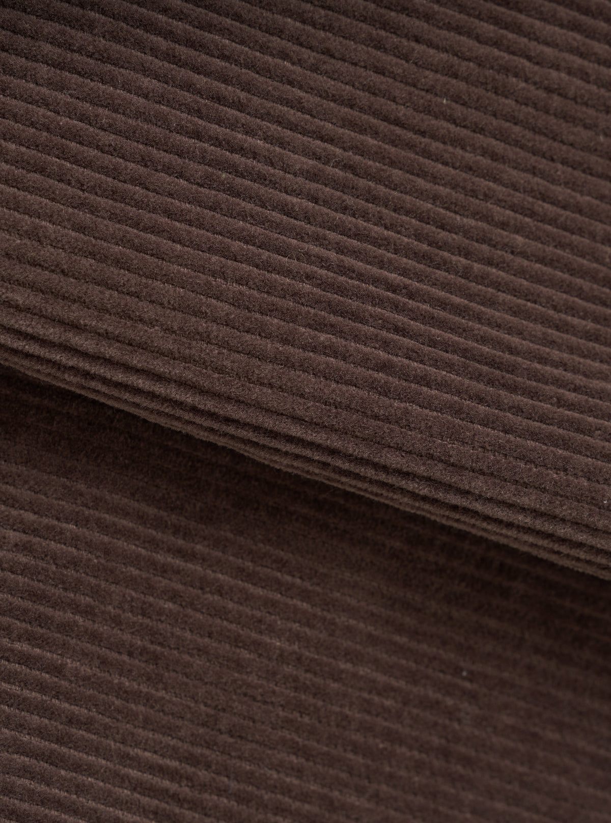 Close-up of the cacao brown corduroy fabric of the utility jacket, showing wide-wale cord texture with soft, dense pile. A folded edge runs diagonally across the frame, revealing the fabric's thickness. No background visible.