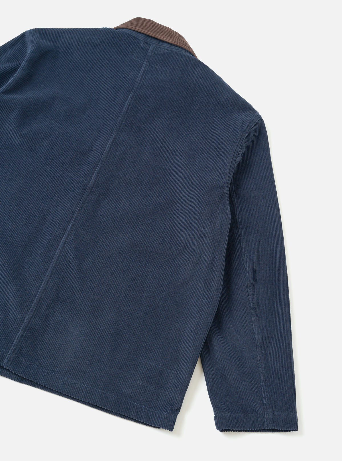 Dark navy corduroy utility jacket, flat lay on a light grey seamless backdrop. Shot shows the back and right sleeve at an angle. Wide-wale cord texture is prominent throughout. A contrast brown corduroy collar is visible at the top. Vertical back seams and a straight hem with visible stitching are shown.