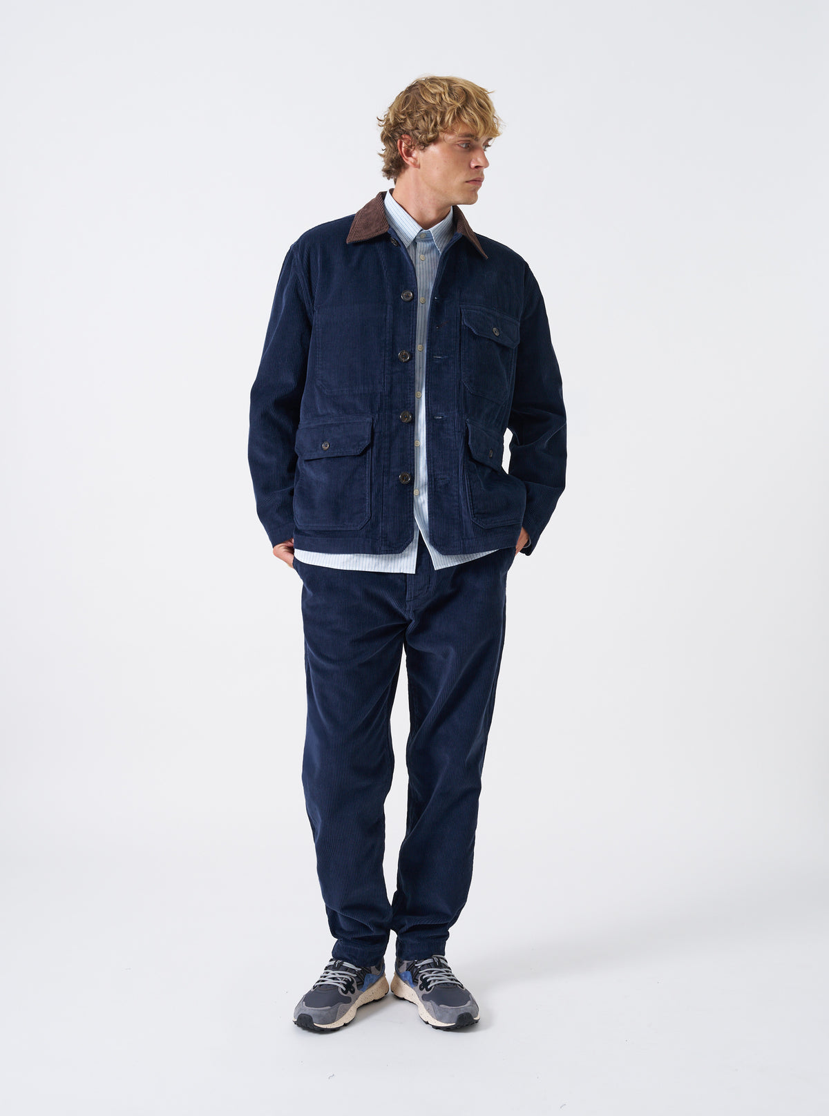 A male model wears the Dark Navy Cord Utility Jacket standing in a full front view on a clean light studio background. The jacket features structured utility pockets, button closures, and fine corduroy weave.