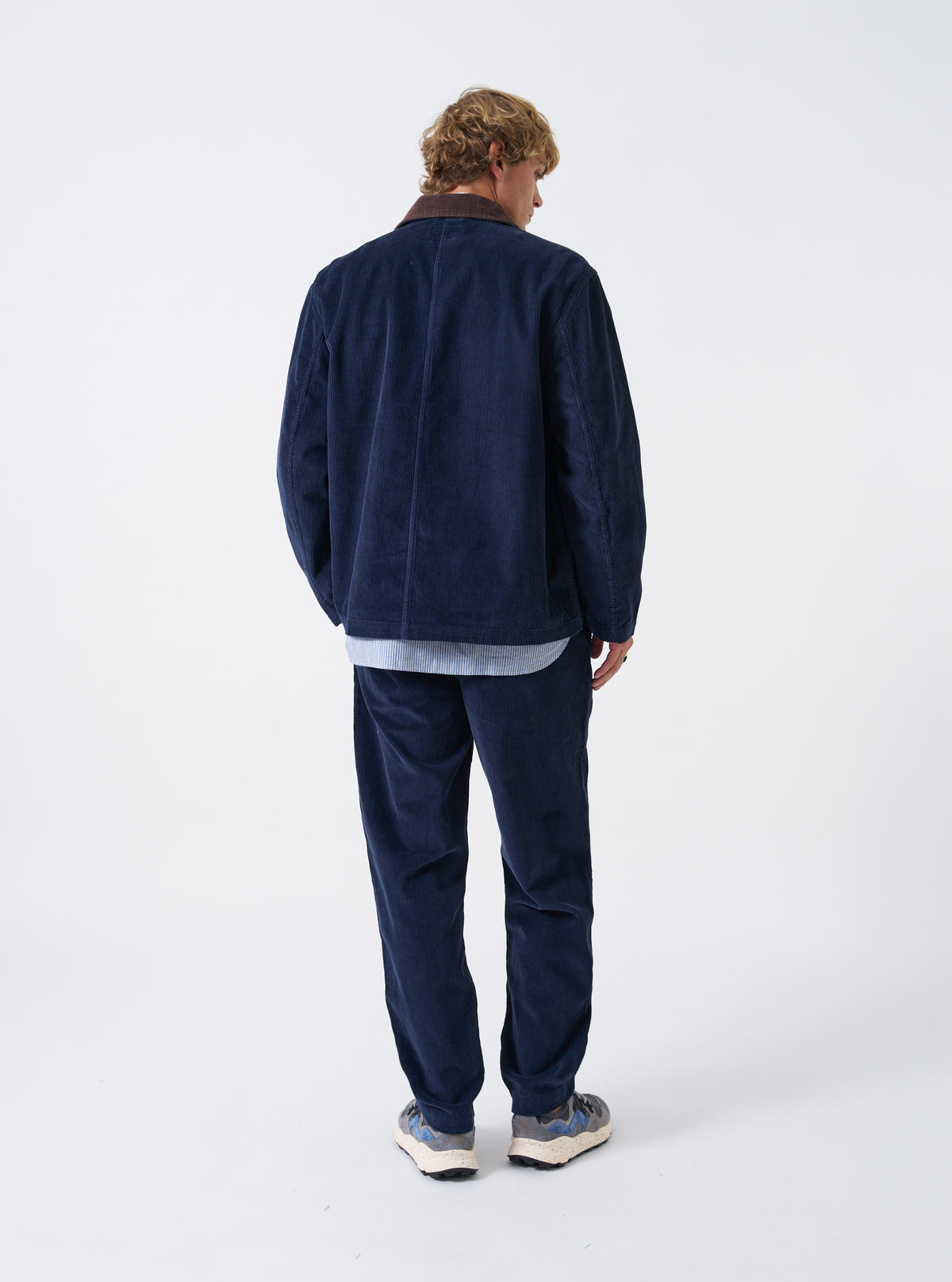 A rear view of a male model wearing the Dark Navy Cord Utility Jacket against a plain off-white studio background. The photo highlights the relaxed tailored cut, hemline, and deep navy corduroy fabric.