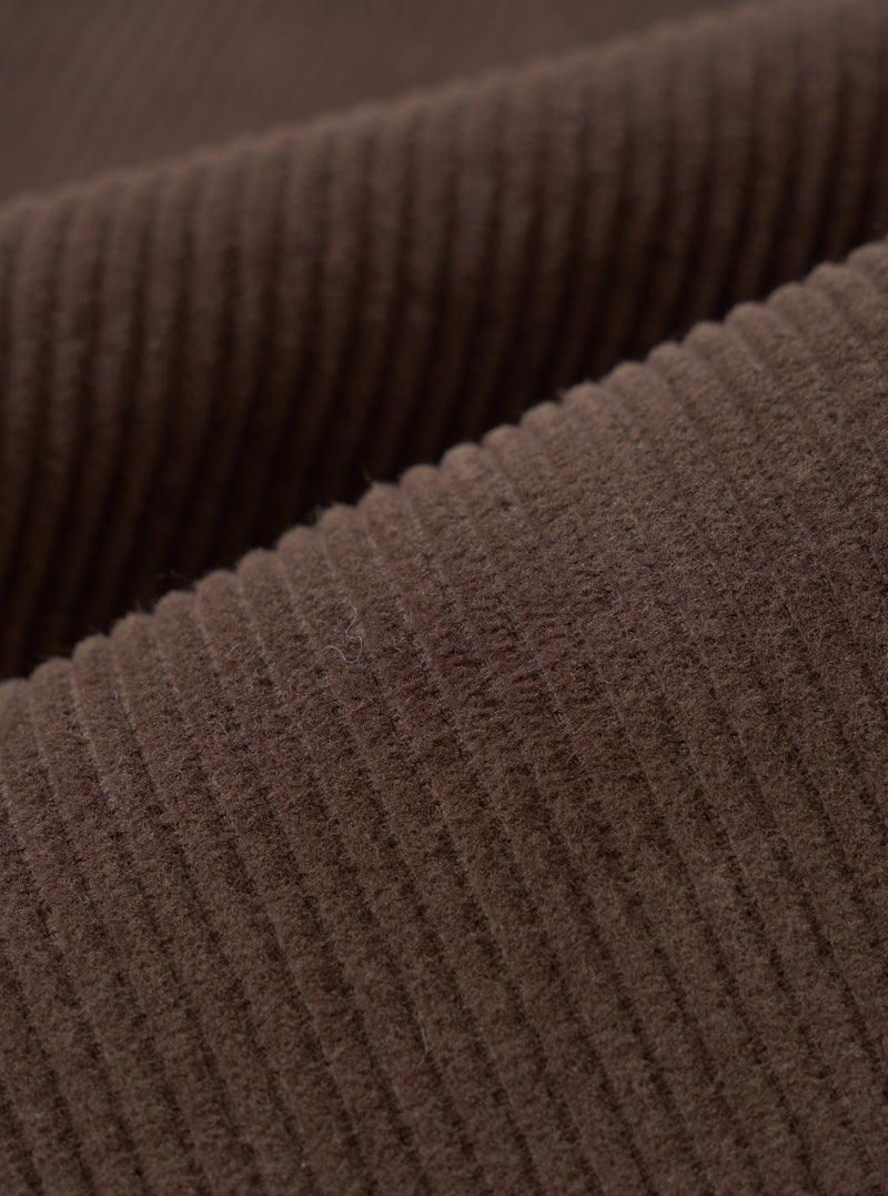 Close-up detail shot of cacao brown corduroy fabric, showing wide, clearly defined wales running diagonally across the frame. The raised ridges and textured valleys of the cord are sharply in focus, highlighting the soft, dense pile and matte finish of the material.