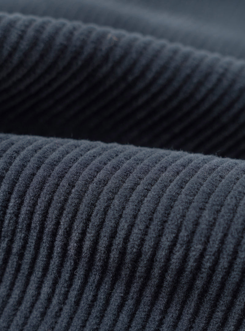 Close-up fabric detail of the dark navy cord military chino, showing wide-wale corduroy texture with raised ridges running diagonally across the frame. Two overlapping layers of the fabric are visible, highlighting the deep navy tone and soft, dense pile of the cord.