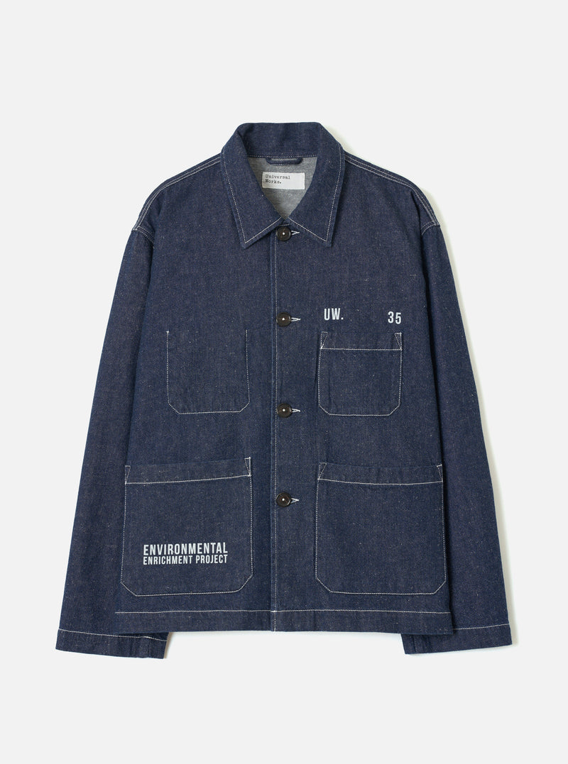 Indigo denim chore jacket laid flat on a light grey seamless backdrop. Point collar, four dark brown buttons down the placket, and contrasting white topstitching throughout. Two chest patch pockets and two large lower patch pockets. White printed text reads "UW. 35" on the chest and "Environmental Enrichment Project" on the lower left pocket. Universal Works label visible at the collar.