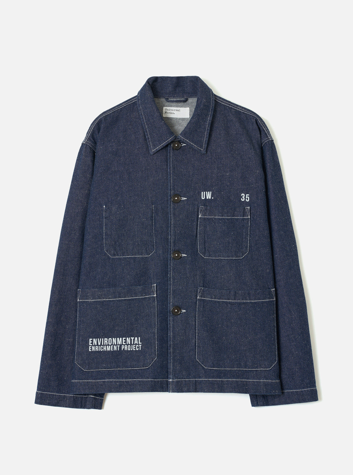 Indigo denim chore jacket laid flat on a light grey seamless backdrop. Point collar, four dark brown buttons down the placket, and contrasting white topstitching throughout. Two chest patch pockets and two large lower patch pockets. White printed text reads "UW. 35" on the chest and "Environmental Enrichment Project" on the lower left pocket. Universal Works label visible at the collar.