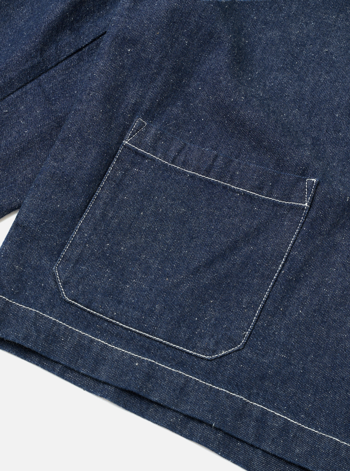 Close-up flat lay of the indigo future denim Ives jacket on a white surface, showing a large patch pocket with contrast white topstitching and angled bartack reinforcement at the corners. The hem and pocket base feature the same white stitching. The slubby denim texture is visible throughout.