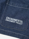 Close-up of the chest patch pocket on an indigo denim jacket, showing white contrast stitching and a printed "Environmental Enrichment Project" slogan in white block capitals. The raw indigo denim has a flecked texture. Shot flat on a white surface.