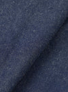 Close-up of the indigo future denim fabric used in the Ives Jacket, showing a mid-blue woven textile with flecks of white and cream throughout. The cloth is folded to reveal texture and depth, with a slightly slubbed, neppy surface typical of denim with natural fibre variation.