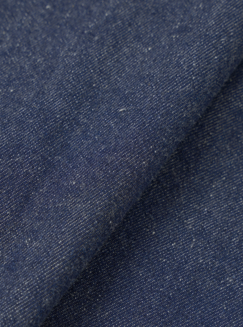 Close-up of the indigo future denim fabric used in the Ives Jacket, showing a mid-blue woven textile with flecks of white and cream throughout. The cloth is folded to reveal texture and depth, with a slightly slubbed, neppy surface typical of denim with natural fibre variation.