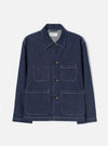 Indigo denim chore jacket laid flat on a light grey studio backdrop. Features a pointed shirt collar, four-button front placket with dark buttons, and four large patch pockets outlined with contrast white topstitching. White stitching details run throughout. A Universal Works woven label sits at the inner collar.