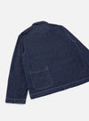 Back view of the indigo denim Ives Jacket, laid flat on a light grey seamless backdrop. Raw indigo denim fabric with visible texture and flecks. White contrast topstitching runs along the seams, hem and cuffs. A large patch pocket sits at the lower back. Small woven label at lower right. Shirt-style collar visible at top.