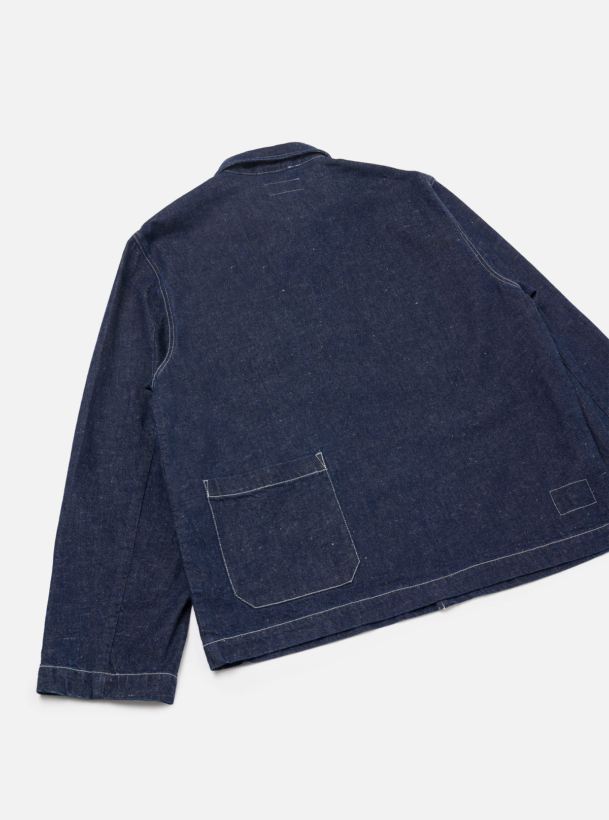 Back view of the indigo denim Ives Jacket, laid flat on a light grey seamless backdrop. Raw indigo denim fabric with visible texture and flecks. White contrast topstitching runs along the seams, hem and cuffs. A large patch pocket sits at the lower back. Small woven label at lower right. Shirt-style collar visible at top.