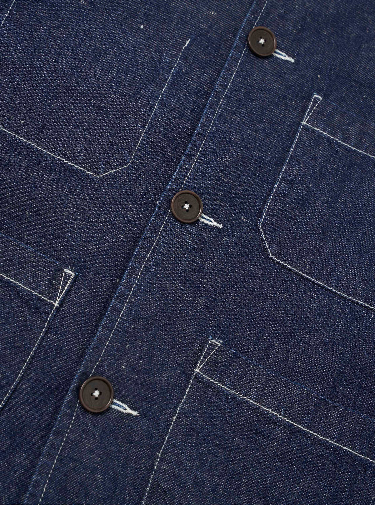Close-up detail of the indigo Future Denim Ives Jacket, showing three dark horn buttons along the front placket and four large patch pockets with contrast white topstitching. The raw indigo denim texture is clearly visible. Shot flat on a plain white background.