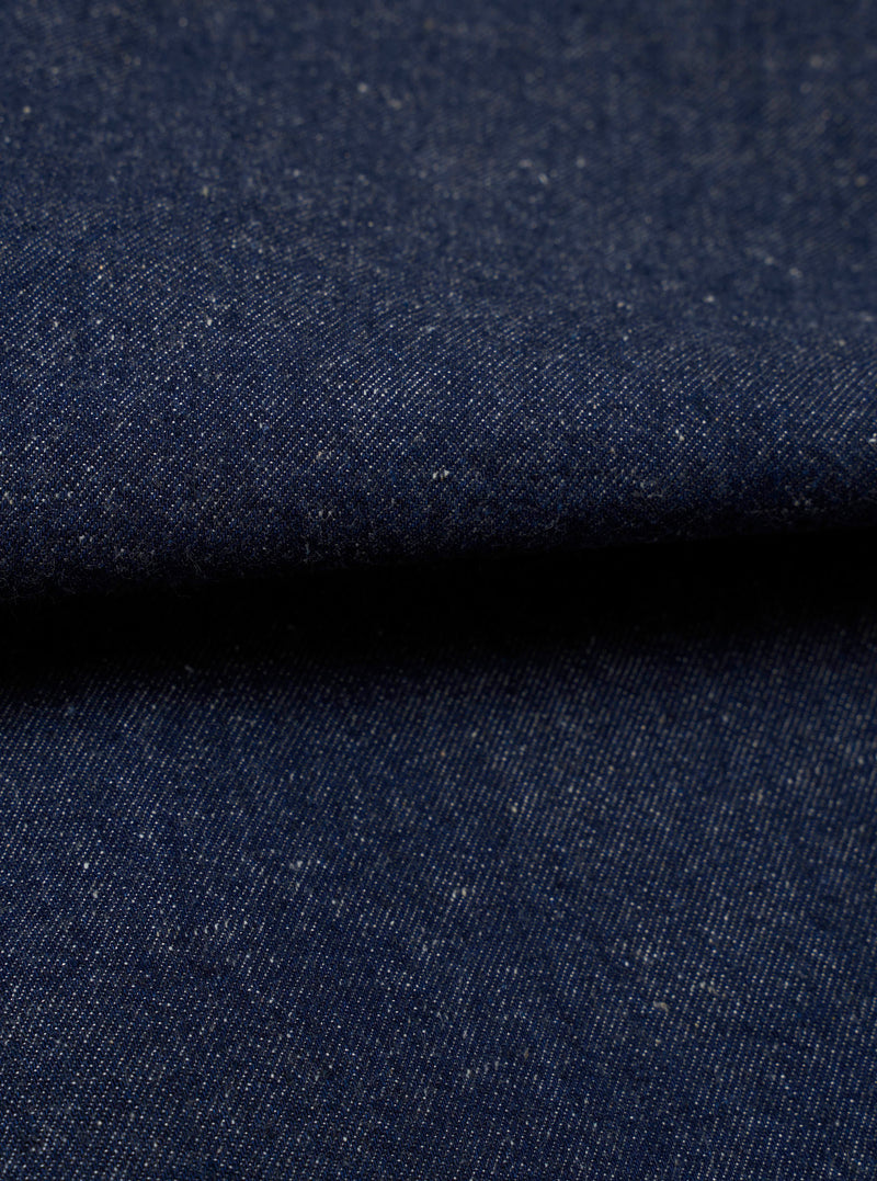 Close-up fabric shot of the Indigo Future Denim Ives Jacket. The deep indigo denim shows a fine twill weave with scattered flecks of white and ecru slub throughout the yarn, giving the fabric a neppy, textured appearance. The fabric is folded, creating a raised ridge across the centre of the frame against a dark background.