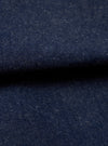 Close-up fabric shot of the Indigo Future Denim Ives Jacket. The deep indigo denim shows a fine twill weave with scattered flecks of white and ecru slub throughout the yarn, giving the fabric a neppy, textured appearance. The fabric is folded, creating a raised ridge across the centre of the frame against a dark background.