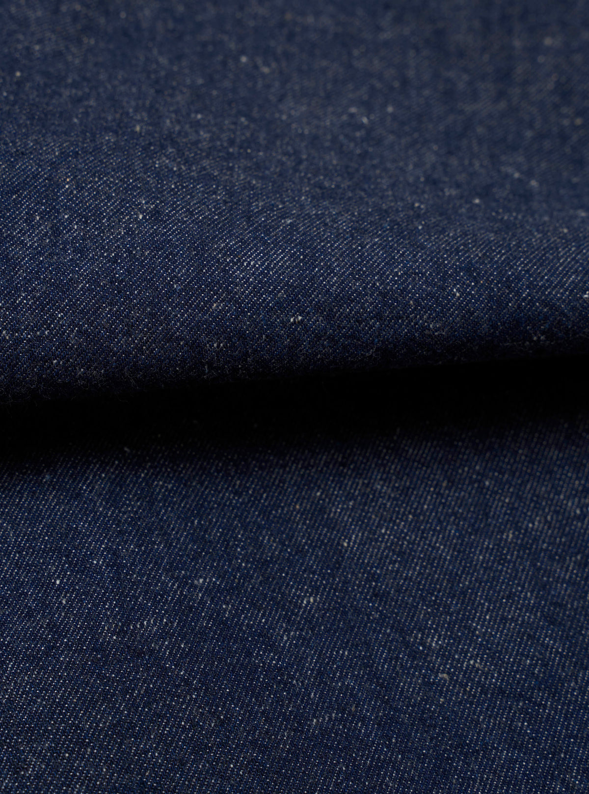 Close-up fabric shot of the Indigo Future Denim Ives Jacket. The deep indigo denim shows a fine twill weave with scattered flecks of white and ecru slub throughout the yarn, giving the fabric a neppy, textured appearance. The fabric is folded, creating a raised ridge across the centre of the frame against a dark background.