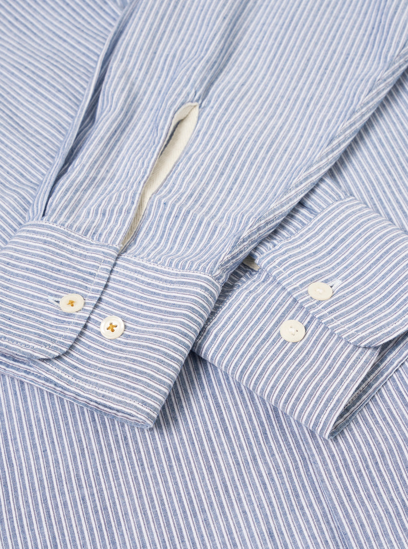 Angled product shot of the Blue Montgomery Stripe Station Shirt against a plain light background. The composition highlights the blue stripe alignment along the side seams, clean buttoned cuffs, crisp collar fold, and relaxed fit.