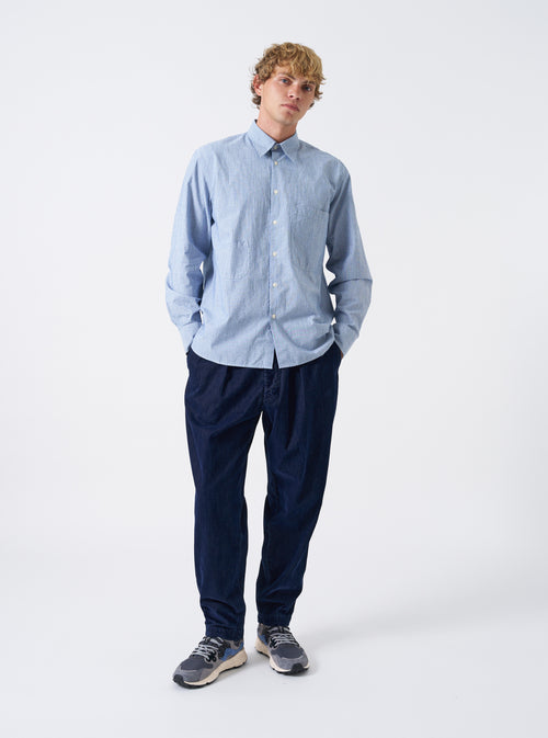 A male model wears the Blue Montgomery Stripe Station Shirt buttoned up, posing against a neutral studio backdrop. The full-length shot illustrates the blue vertical stripe pattern, classic collar, chest pockets, and comfortable relaxed fit.