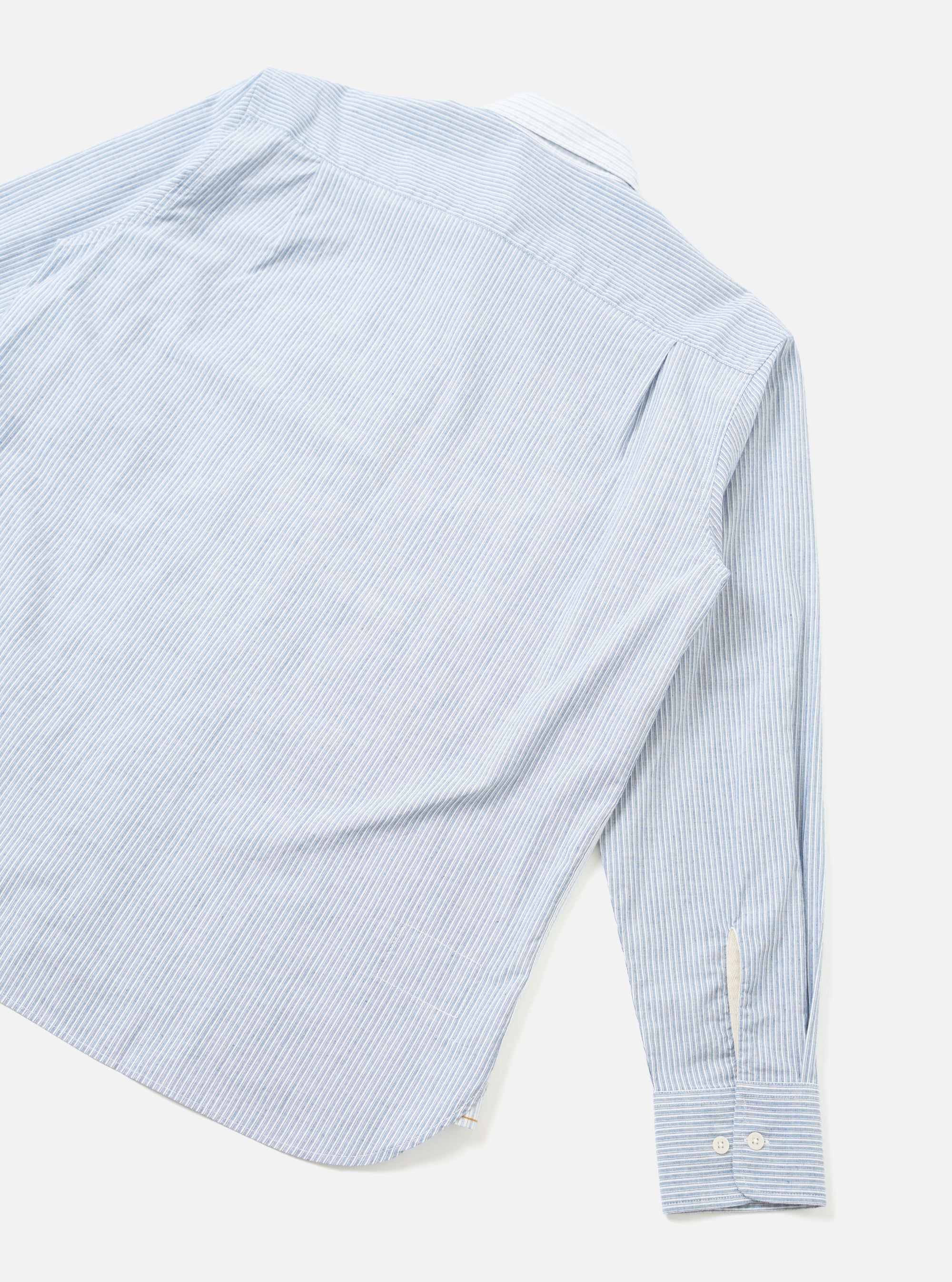 Flat lay of a light blue and white mix-stripe station shirt on a white studio surface. The shot crops the upper-left corner, showing the back panel, one long sleeve, and a rounded hem. The cuff is buttoned with two small white buttons and features a barrel-style finish. A small woven label is visible at the hem seam.