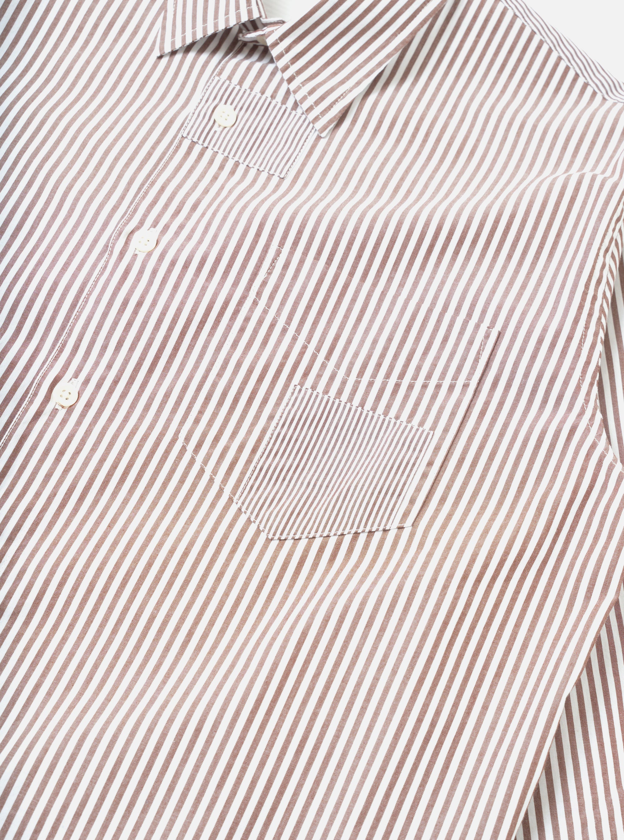 Close-up flat lay of a brown and white striped cotton shirt on a white background. The point collar, button placket with white buttons, and a chest patch pocket are visible. The pocket features a contrasting finer stripe panel. Topstitching outlines the pocket and placket seams.
