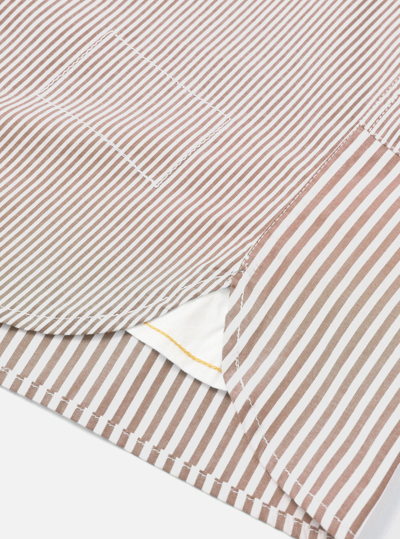 Close-up detail of a brown and white striped cotton shirt laid flat on a white surface. The shot focuses on the curved shirt tail hem, a chest patch pocket, and side seam stitching. A white inner label with a gold detail is visible at the hem opening.