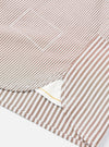 Close-up detail of a brown and white striped cotton shirt laid flat on a white surface. The shot focuses on the curved shirt tail hem, a chest patch pocket, and side seam stitching. A white inner label with a gold detail is visible at the hem opening.